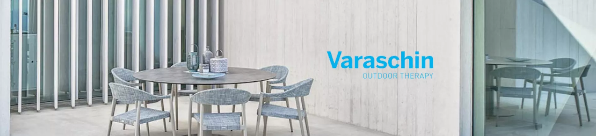 Varaschin Outdoor Therapy, runder Tisch mit grauen Stühlen auf einer Terrasse vor einer modernen, weißen Wand.
