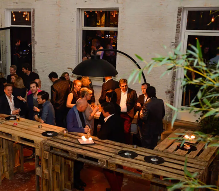 Heatsail Dome Arc Stehlampe in Schwarz beleuchtet eine gesellige Abendveranstaltung im Freien mit rustikalen Holztischen.