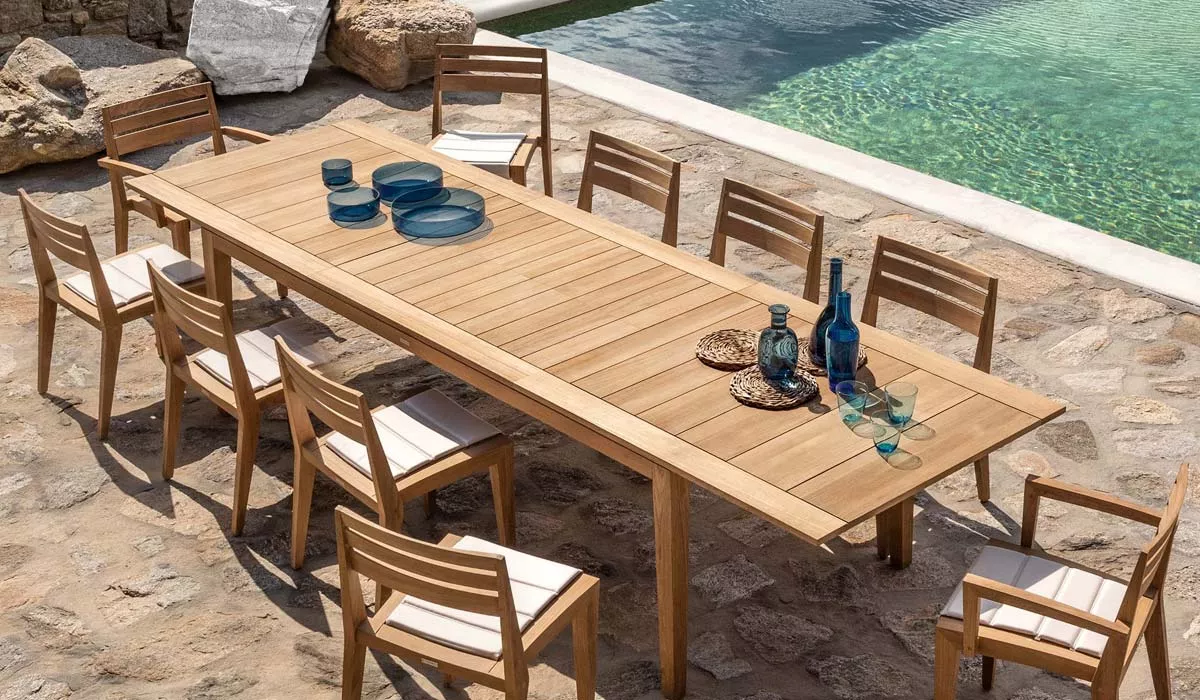 Ethimo-Gartentisch aus Holz in Naturfarbe, umgeben von passenden Stühlen, auf einer Steinterrasse neben einem Pool, in sommerlicher Atmosphäre.