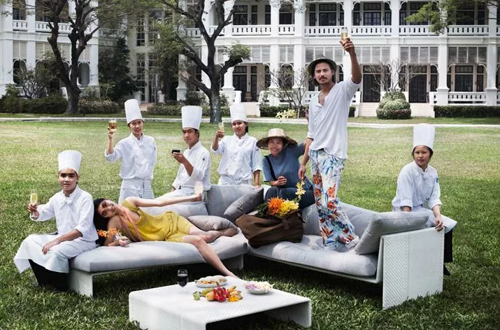 Eine Gruppe von Menschen genießt Getränke auf Dedon Slim Line Gartenmöbeln in einem Garten vor einem großen weißen Gebäude.