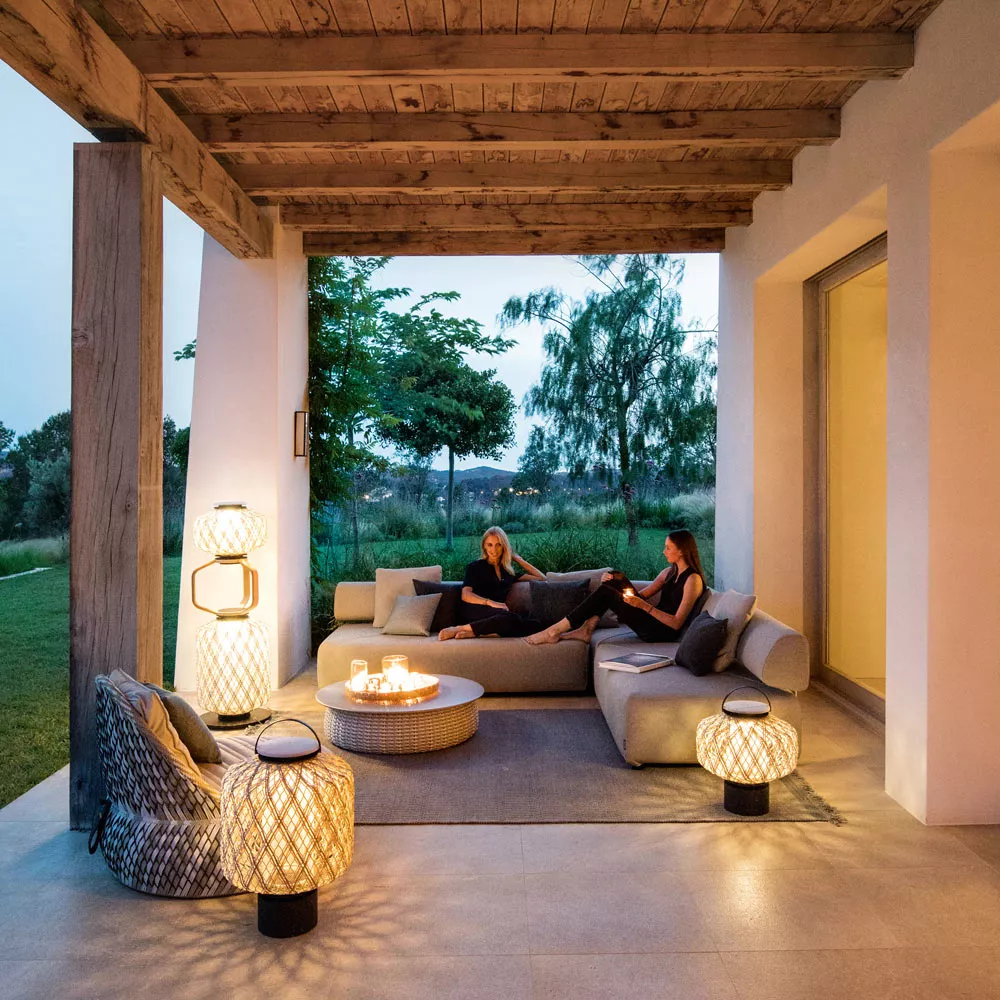 Zwei Frauen sitzen auf einem Sofa in einer überdachten Terrasse umgeben von Dedon The Others Gartenleuchten und Laternen bei Dämmerung.