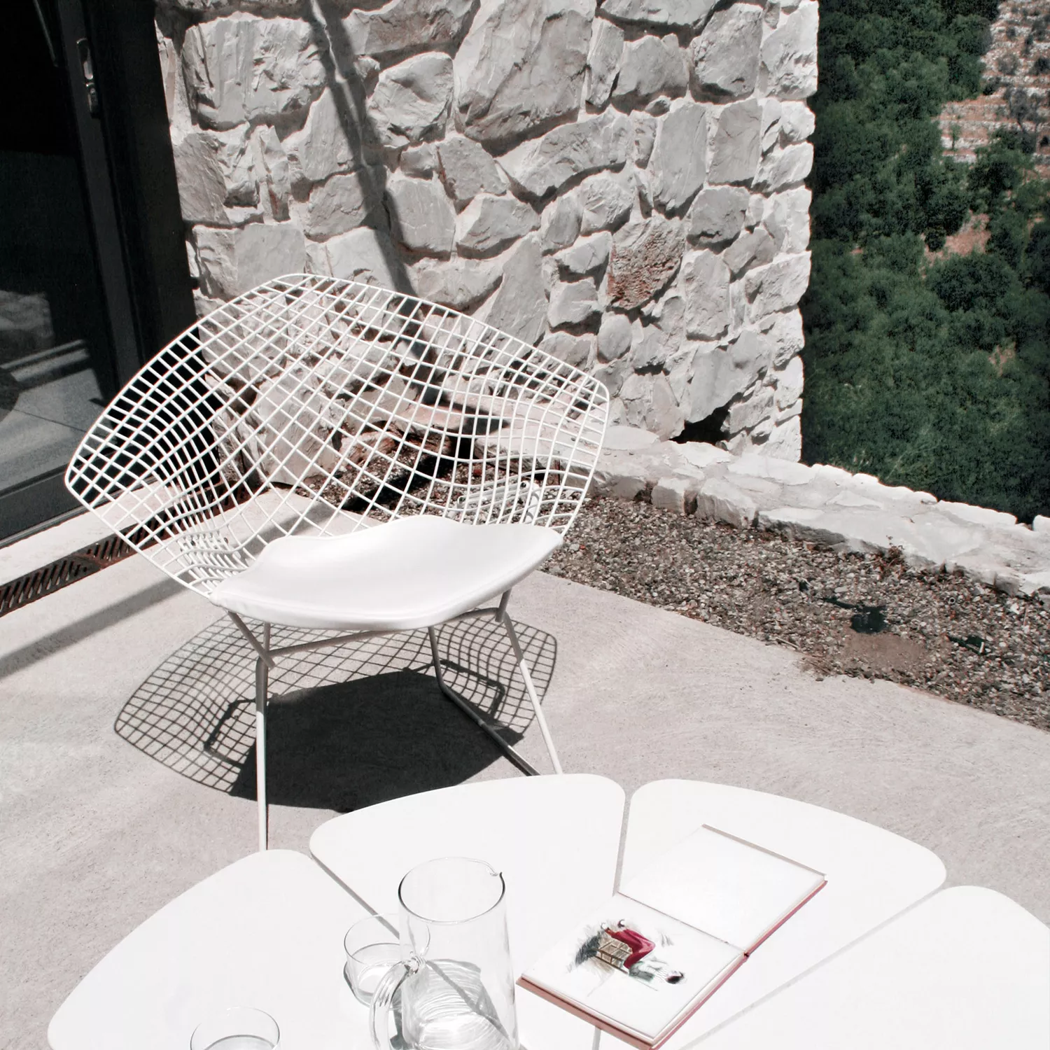Knoll Harry Bertoia Diamond Chair in Weiß auf einer Terrasse mit Steinmauer im Hintergrund, sommerliche Stimmung.
