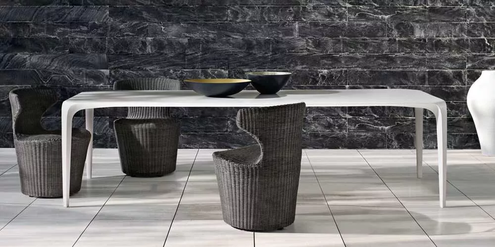 B&B Italia Link Outdoor Essgruppe bestehend aus einem weißen Tisch und vier geflochtenen Stühlen auf einer Terrasse vor einer Steinwand.