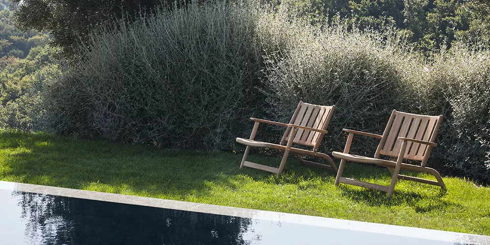 Zwei Roda Levante Holzstühle in natürlicher Umgebung neben einem Pool, umgeben von grüner Vegetation und Büschen.