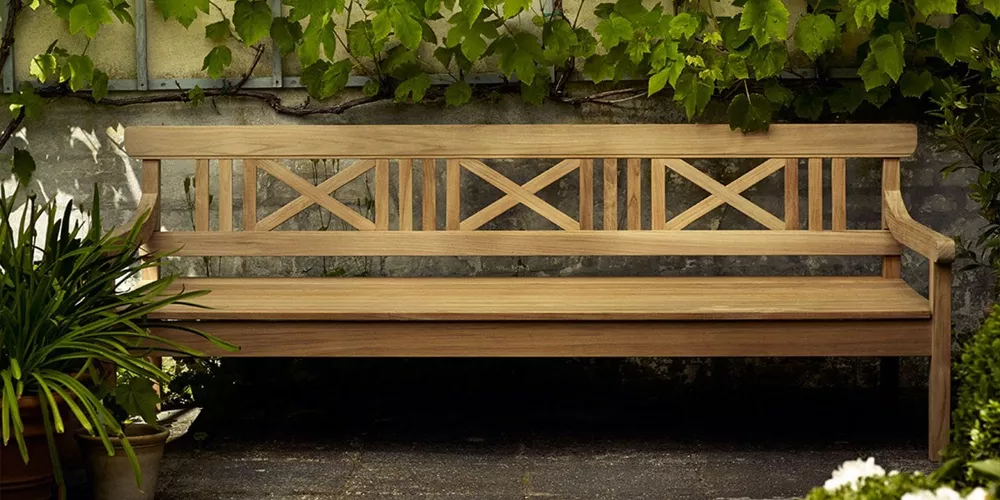 Skagerak Drachmann Bank aus Teakholz, naturfarben, auf einer Terrasse mit Pflanzen im Hintergrund, stimmungsvolles Gartenbild.