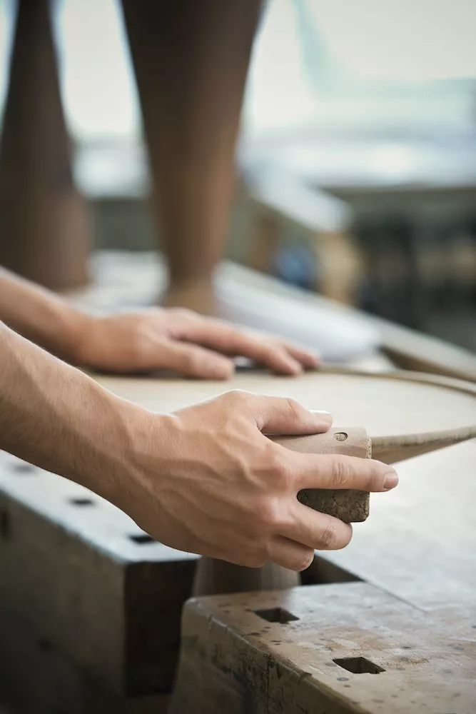 Zwei Hände bearbeiten ein Holzstück in einer Werkstatt im Hintergrund sind unscharfe Werkzeuge und Möbel zu sehen