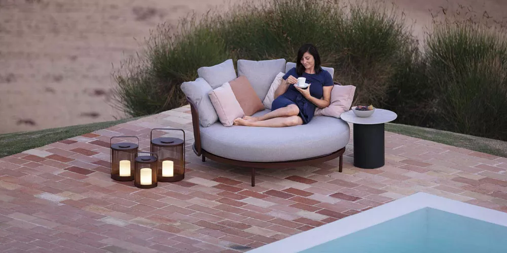 Frau entspannt auf grauem Todus Loungesessel neben Pool, umgeben von Laternen auf Ziegelsteinterrasse, bei Abenddämmerung.