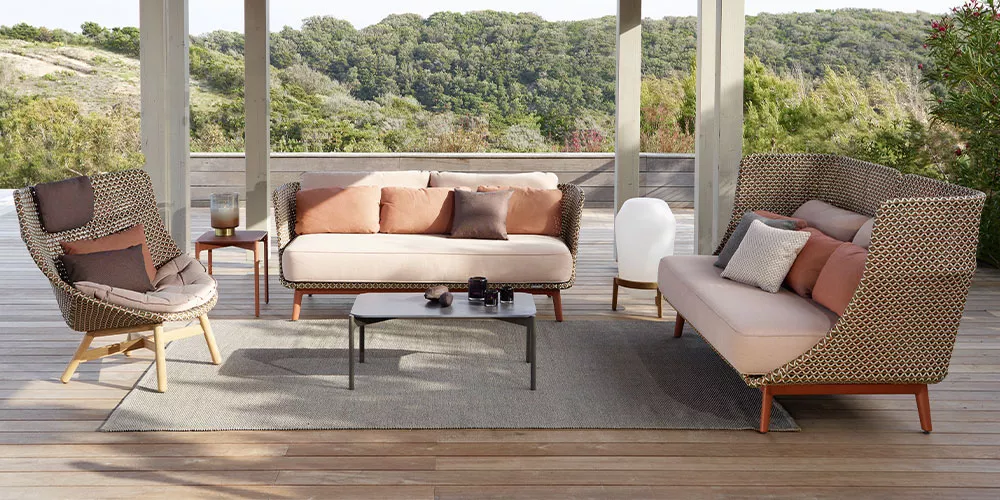 Dedon Loungesofa Mbraq auf einer Terrasse mit Kissen und einem Tisch in einer natürlichen Umgebung mit Bäumen im Hintergrund