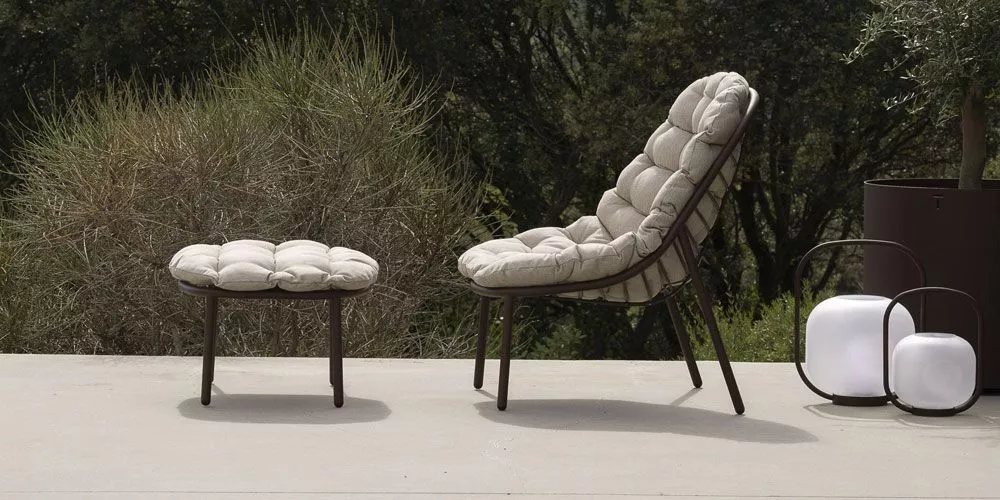 Todus Albus Kollektion in Beige auf einer Terrasse mit Bäumen im Hintergrund, bestehend aus einem gepolsterten Stuhl und Hocker.