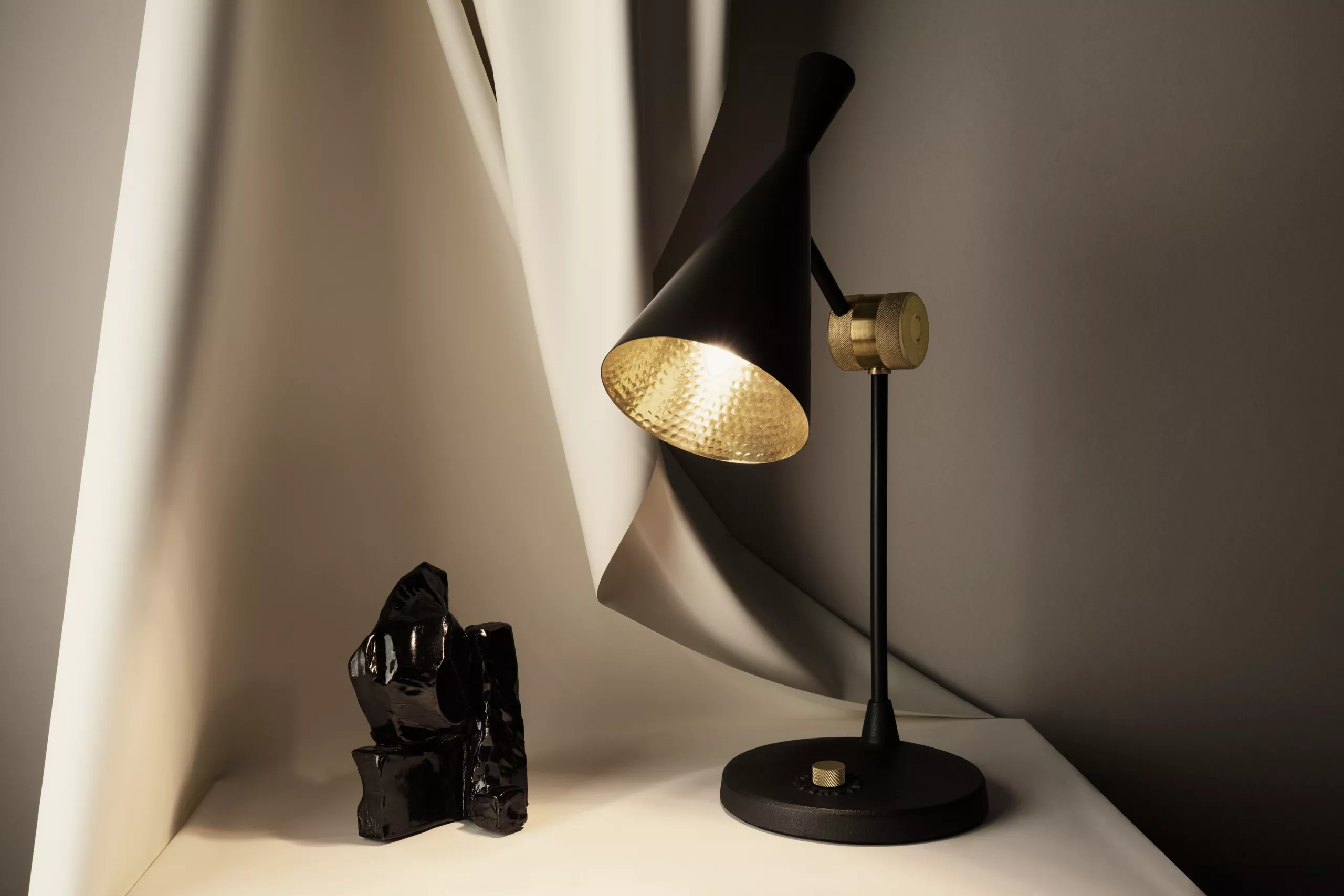 Tom Dixon Beat LED Tischleuchte in Schwarz auf weißem Tisch vor grauem Hintergrund, stimmungsvolle Beleuchtung.