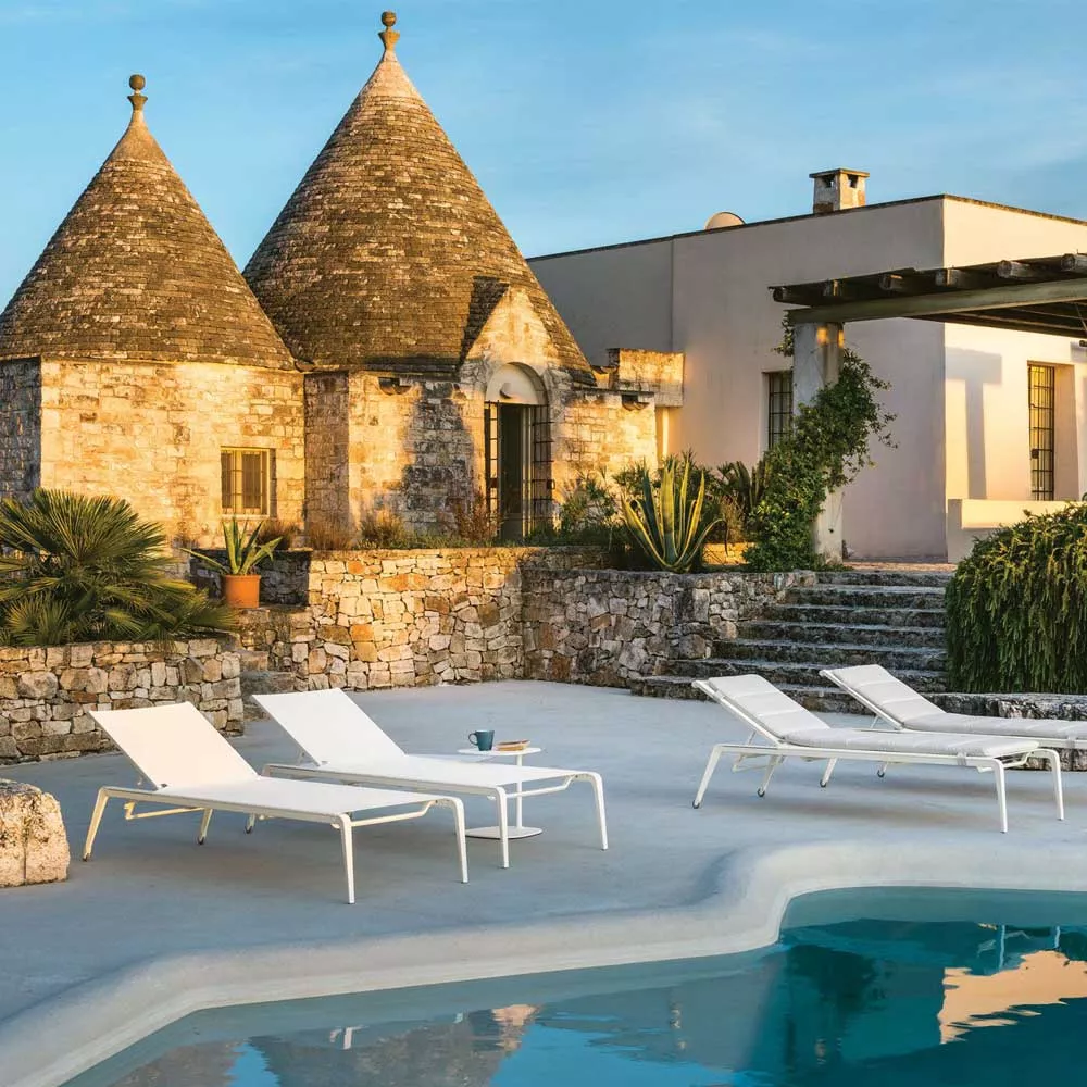 Weiße Fast Ushuaia Sonnenliege auf einer Terrasse mit Pool und historischem Gebäude im Hintergrund, stimmungsvolle Abendbeleuchtung.