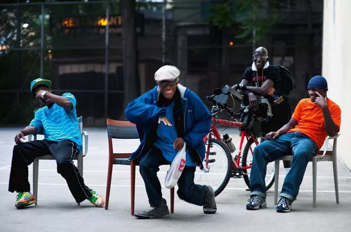 Vier Männer sitzen und tanzen auf Dedon Play Essstühlen im Freien vor einem Gebäude mit einem Fahrrad im Hintergrund.