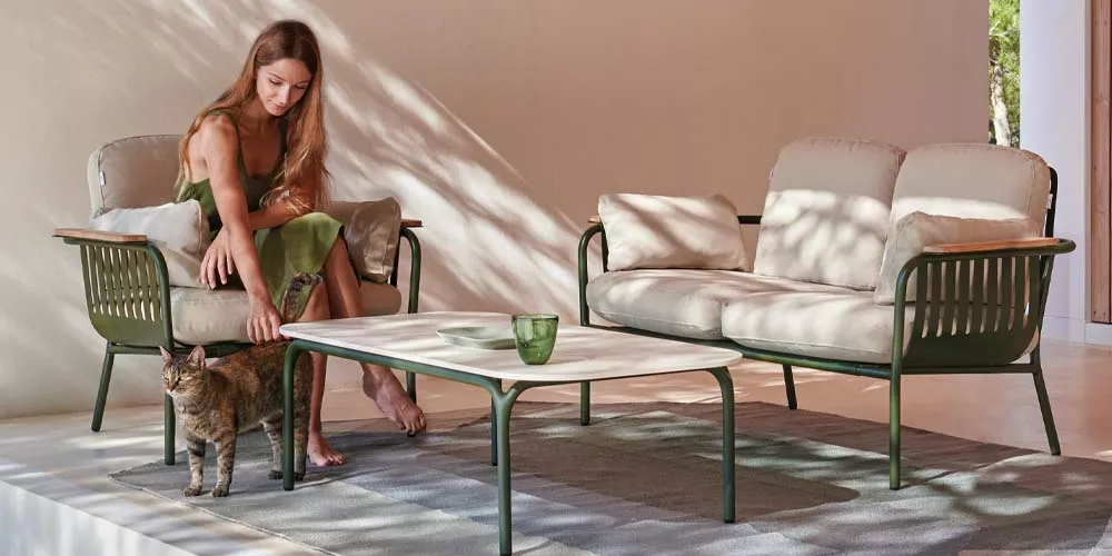 Frau in grünem Kleid streichelt Katze neben Gandia Blasco Capa Lounge-Set in Beige und Grün auf Terrasse mit Schattenmuster.