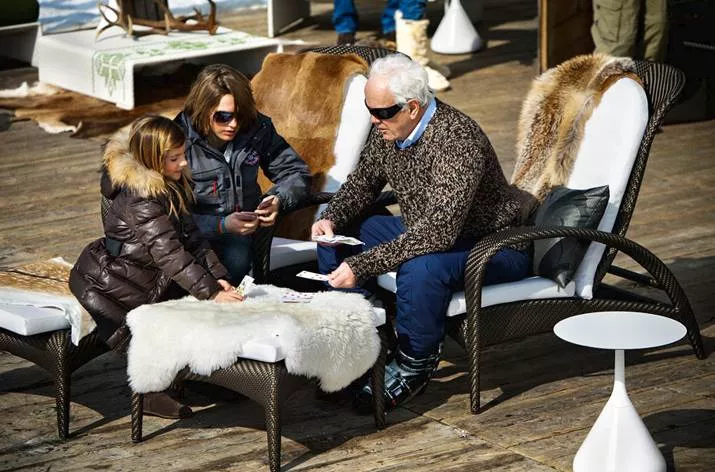 Drei Personen spielen Karten auf einer Terrasse mit Dedon Satellite Esstisch rund 40 cm im Hintergrund und Pelzdecken auf den Stühlen.