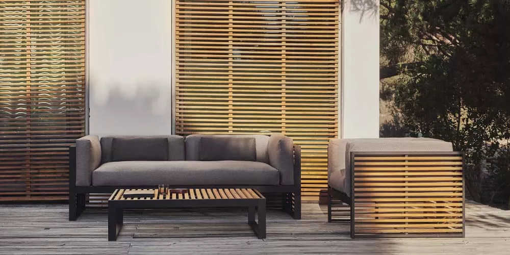 Gandia Blasco DNA Teak Lounge-Set in modernem Design auf einer Holzterrasse vor einer Wand mit Holzjalousien, stimmungsvolles Ambiente.