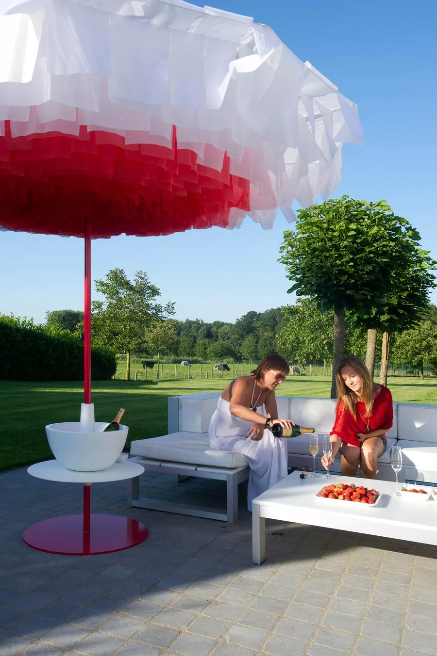 Zwei Frauen sitzen unter einem rot-weißen Sywawa Sonnenschirm Bloom 300 cm auf einer Terrasse und genießen Wein und Erdbeeren.