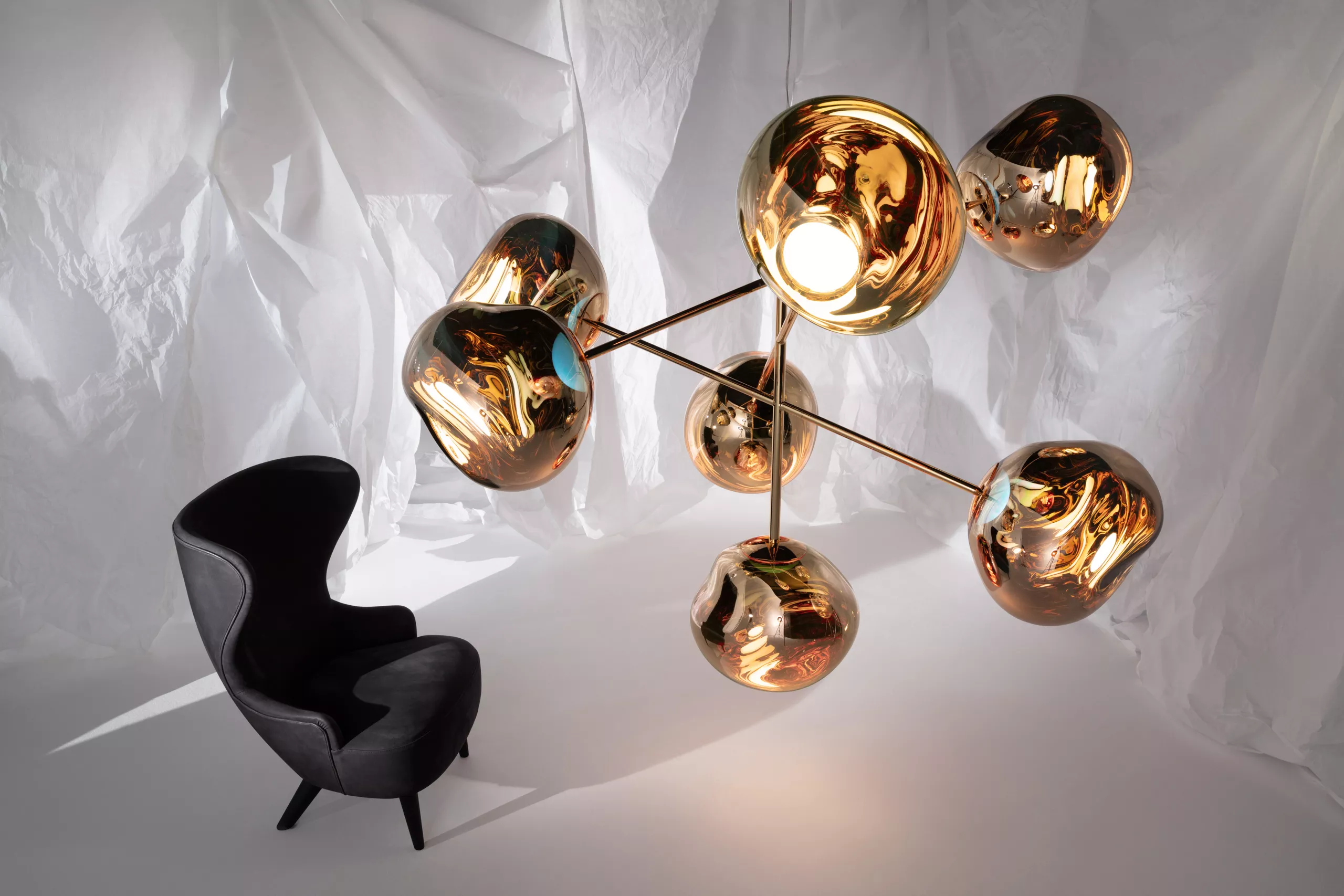 Tom Dixon Kronleuchter MELT in Gold schwebt vor einem weißen Hintergrund neben einem schwarzen Sessel.