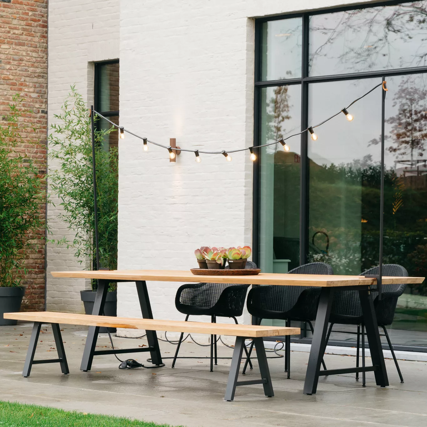 Vincent Sheppard Matteo Bank in hellem Holz vor einer modernen Backsteinfassade mit Lichterkette und Gartenblick.