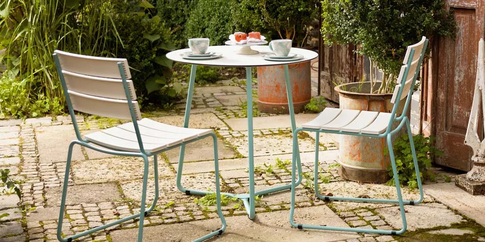 Weißhäupl Balkonmöbel-Set in Weiß und Hellblau auf einer gepflasterten Terrasse mit grüner Pflanzenkulisse.