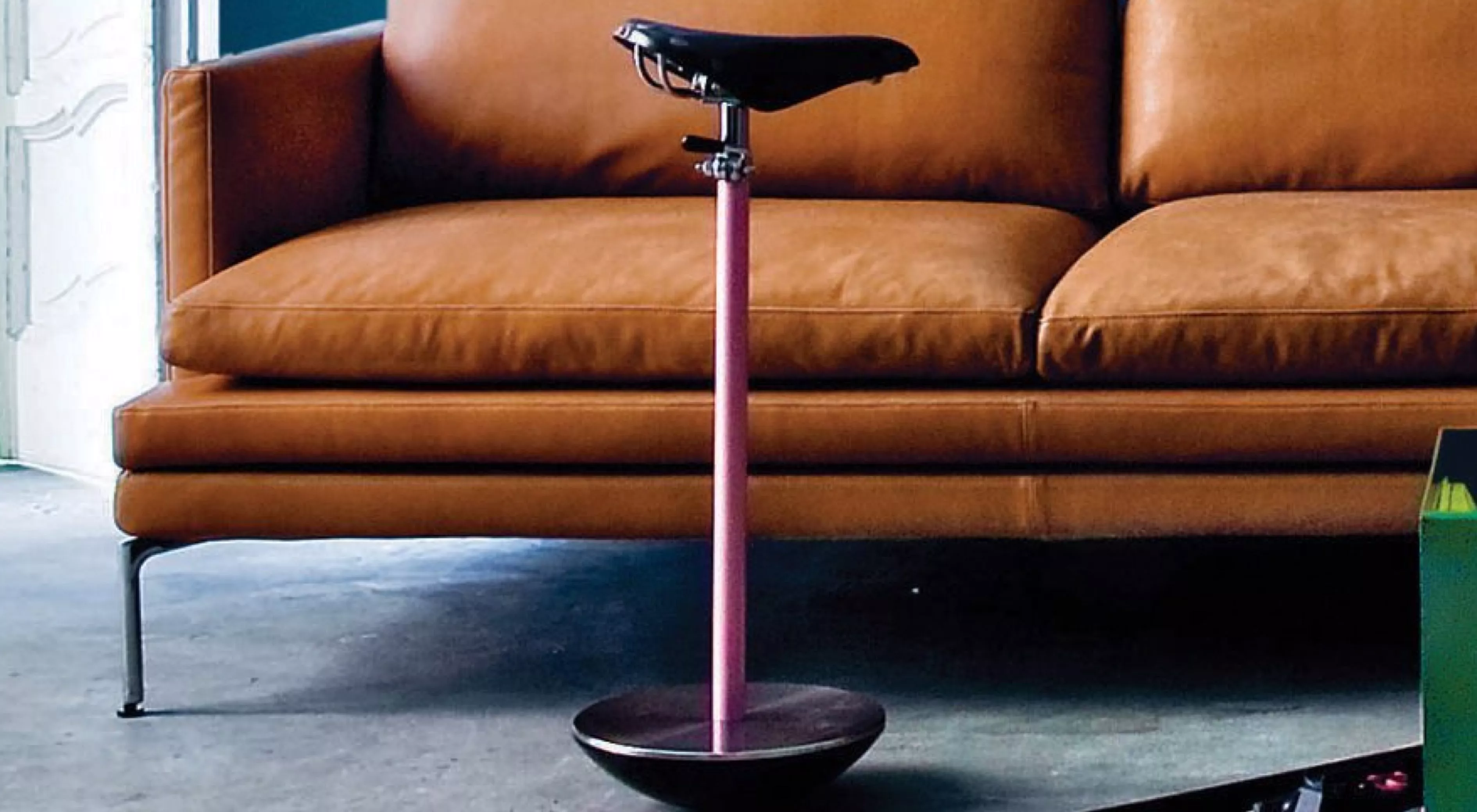 Sella Hocker von Zanotta in Rosa vor einem braunen Ledersofa auf Betonboden, technische Produktpräsentation.