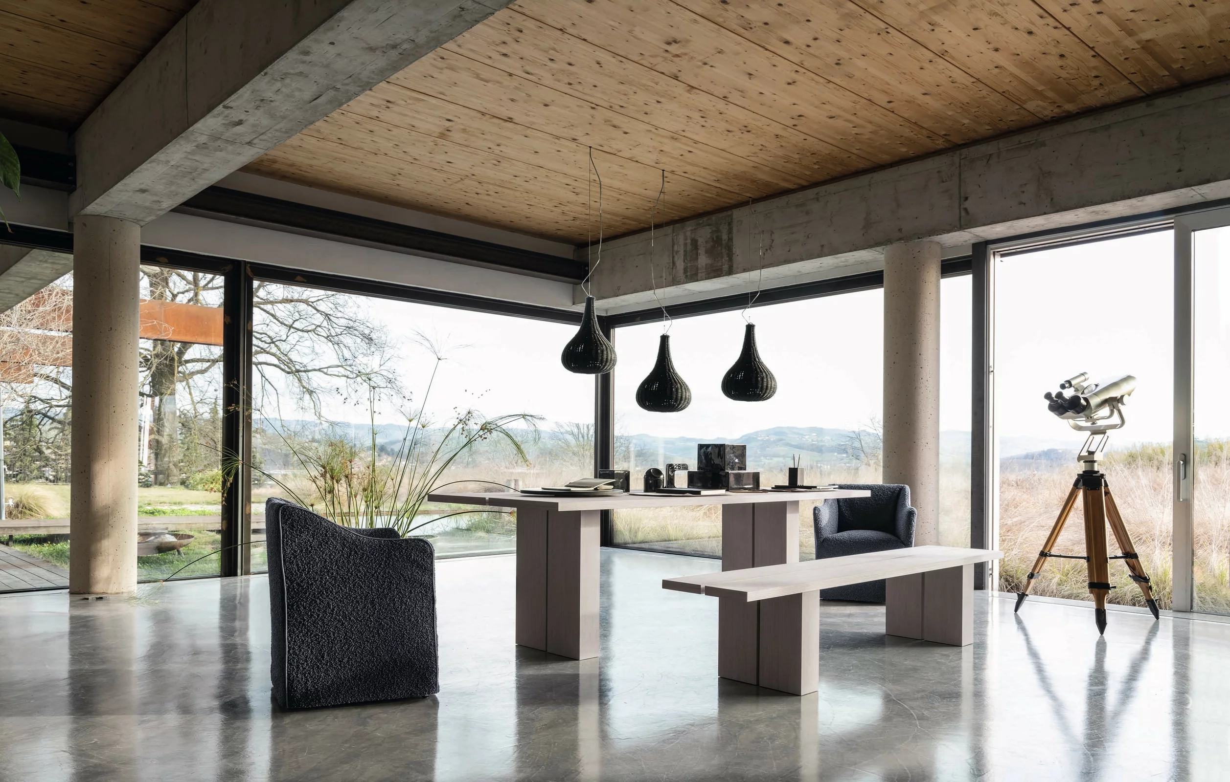 Ein modernes Wohnzimmer mit Gervasoni Tisch und Bank sowie einem Sessel und Teleskop vor großen Fenstern mit Blick auf die Landschaft.