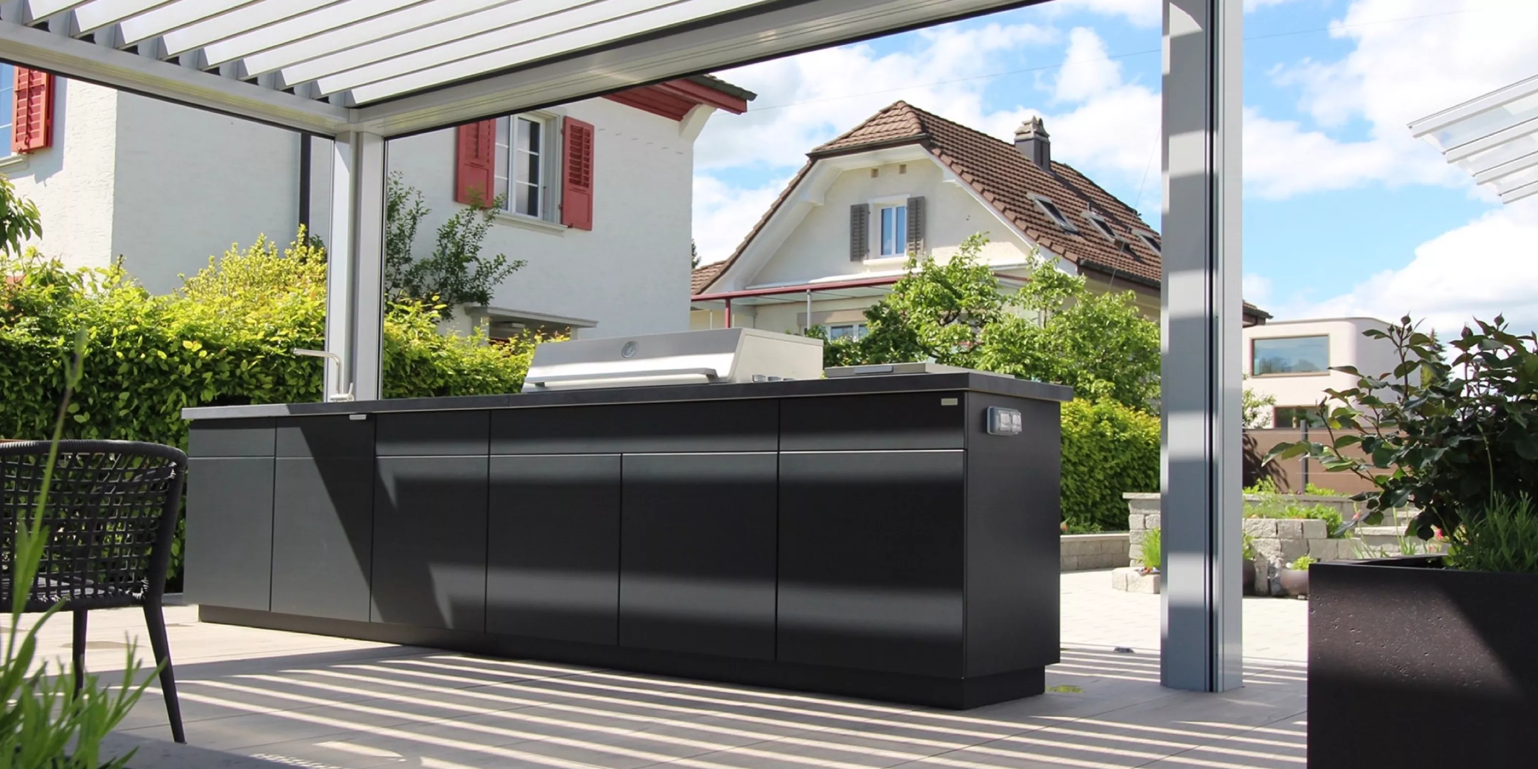 Cubic Outdoor Kitchen in Anthrazit auf einer Terrasse mit modernem Lamellendach und Blick auf ein Haus im Hintergrund.