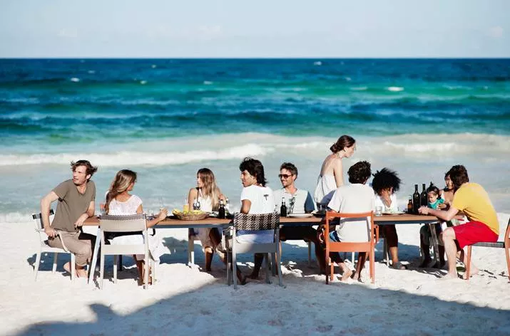 Menschen sitzen an einem Tisch am Strand und genießen eine Mahlzeit mit Blick auf das Meer Dedon Play Essstuhl Dedon Play Outdoor Gartenmöbel
