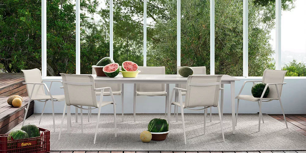 Dedon Kollektion Wa zeigt einen stilvollen Esstisch mit sechs Stühlen in einem modernen Garten mit Wassermelonen auf dem Tisch und im Hintergrund.