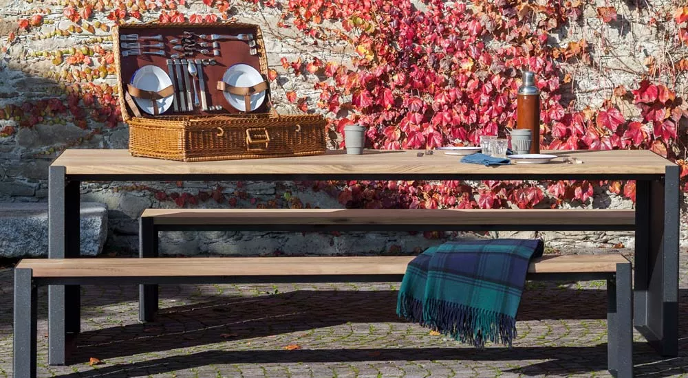 Lapalma PANCO Outdoor-Essgruppe in Holzoptik mit schwarzem Metallgestell, Picknick-Szene vor einer mit roten Blättern bedeckten Steinmauer.