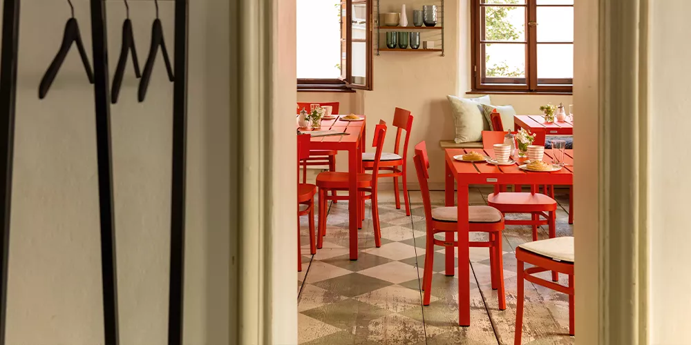 Blick in ein gemütliches Café mit roten Weishäupl Flow Stühlen und Tischen, umgeben von hellen Wänden und großen Fenstern.