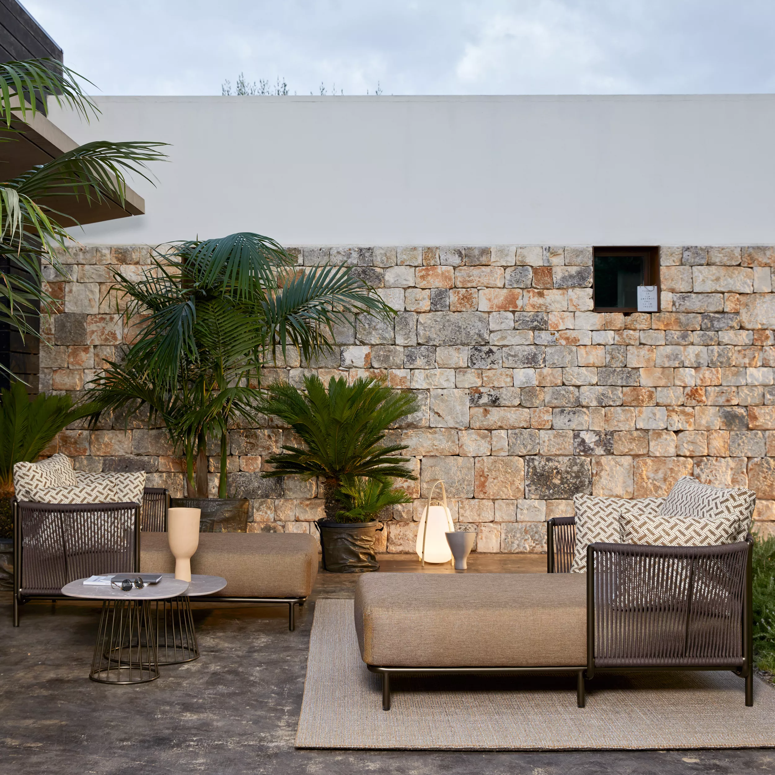 Braunes Roberti Queen Loungemodul vor einer Steinmauer im Garten, umgeben von Pflanzen, mit modernem, gemütlichem Ambiente.