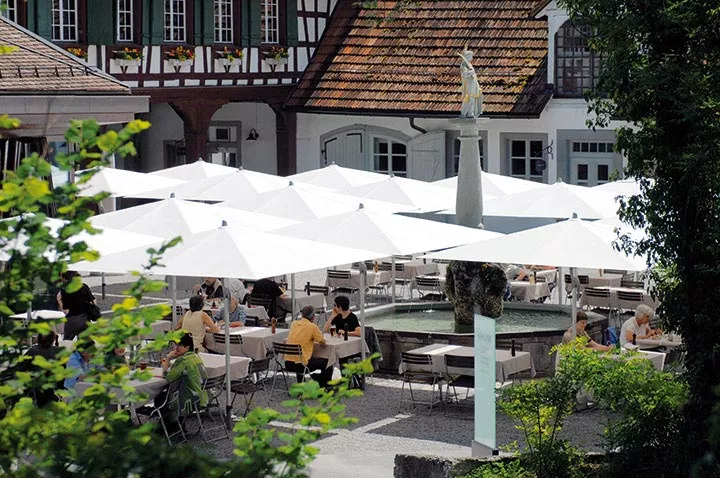 Sonnenschirm Fortello Easy von Glatz in Weiß, aufgestellt in einem belebten Café-Hof mit historischen Gebäuden im Hintergrund.