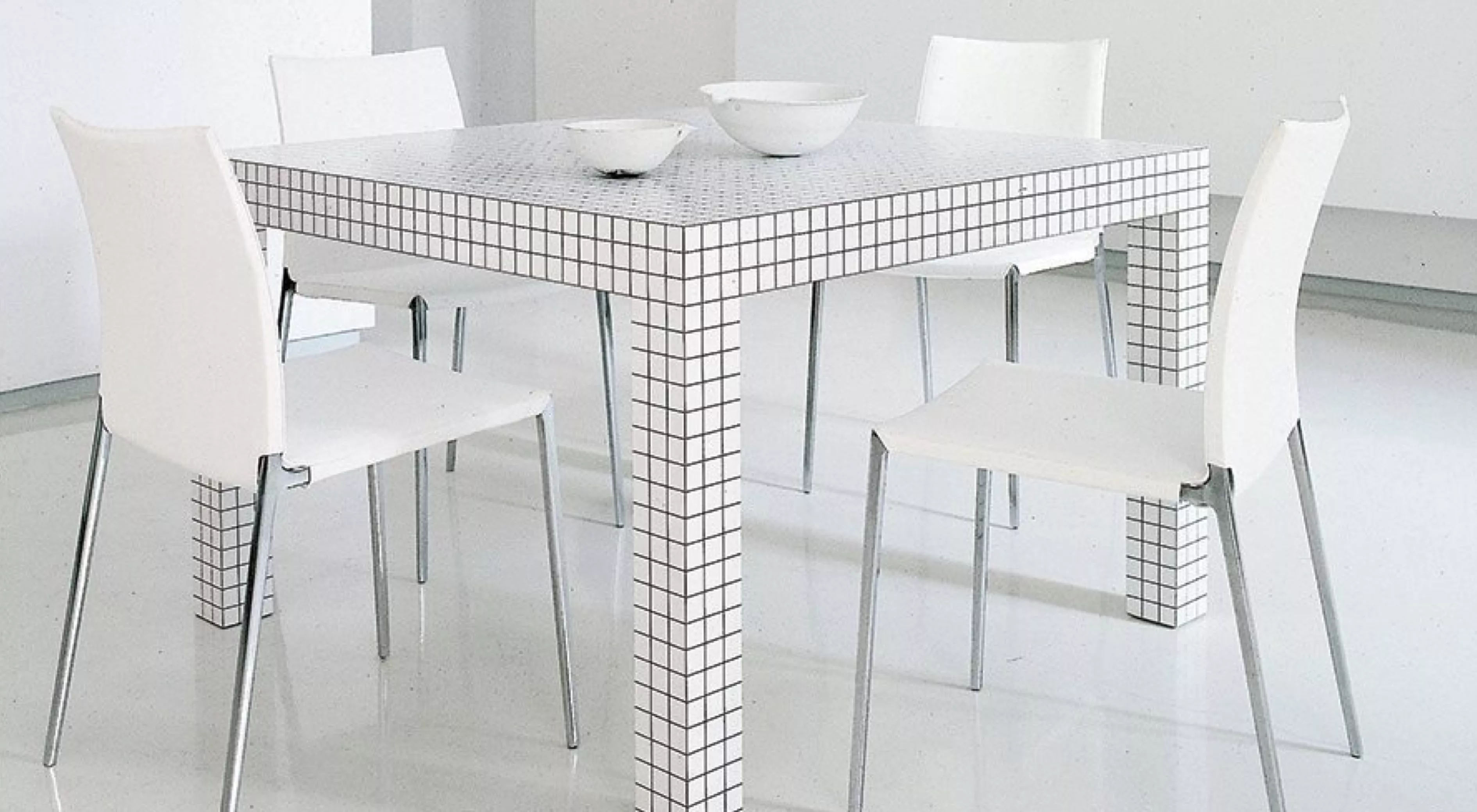 Zanotta Quaderna Tisch in Weiß mit schwarzem Rastermuster, umgeben von vier weißen Stühlen, auf glänzendem Boden in modernem Ambiente.