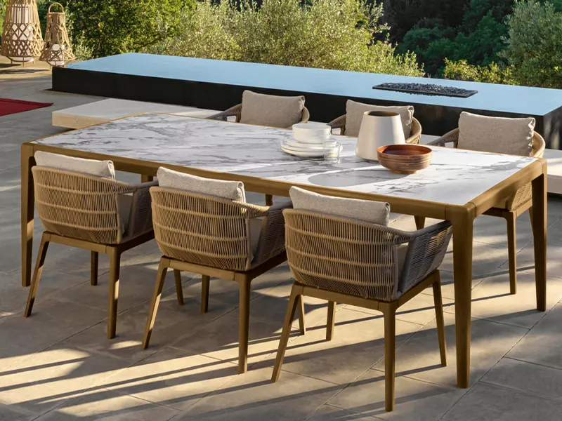 Talenti Cruise Teak Essstuhl in natürlicher Umgebung, mit beigem Polster und Teakholzrahmen, auf einer Terrasse mit grünem Hintergrund.