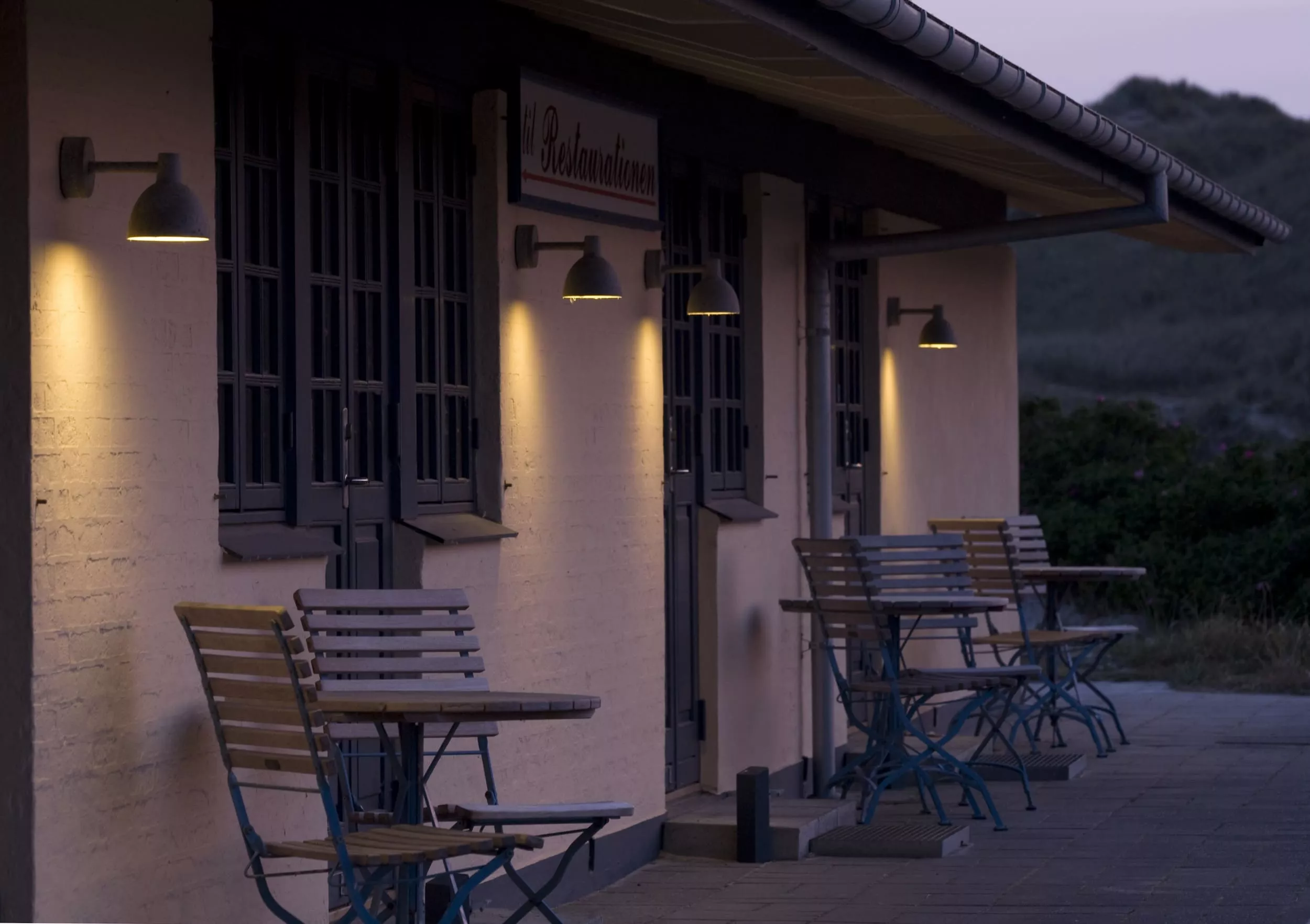 Außenansicht eines Cafés bei Dämmerung mit Holztischen und -stühlen auf einer Terrasse, beleuchtet von Wandlampen.