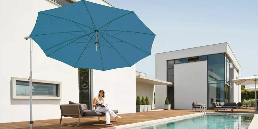 Frau entspannt auf Terrasse unter blauem May Sonnenschirm Dacapo neben modernem Haus und Pool, sonniger Tag, entspannte Stimmung.