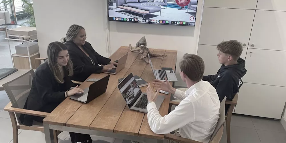 Vier Personen arbeiten an Laptops an einem Holztisch in einem modernen Büro mit einem großen Bildschirm an der Wand.