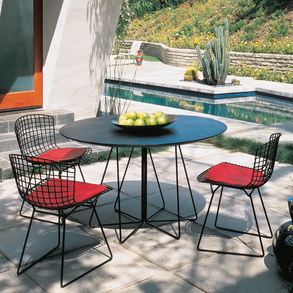 Knoll Paperclip Outdoor Tisch mit Glasplatte und vier Stühlen mit roten Kissen auf einer Terrasse neben einem Pool und Pflanzen im Hintergrund