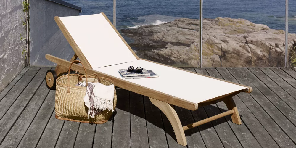 Skagerak Columbus Sonnenliege aus Teakholz mit weißem Stoffbezug auf einer Terrasse mit Meerblick, neben einem Korb mit Handtuch.