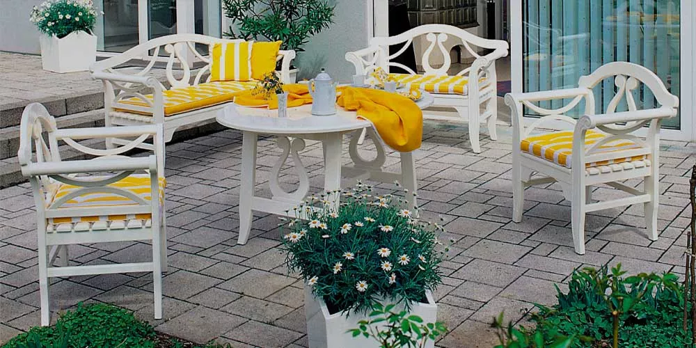 Herrenhaus Michelle Essgruppe in Weiß mit gelben Kissen auf einer Terrasse, umgeben von Pflanzen, bei Tageslicht.