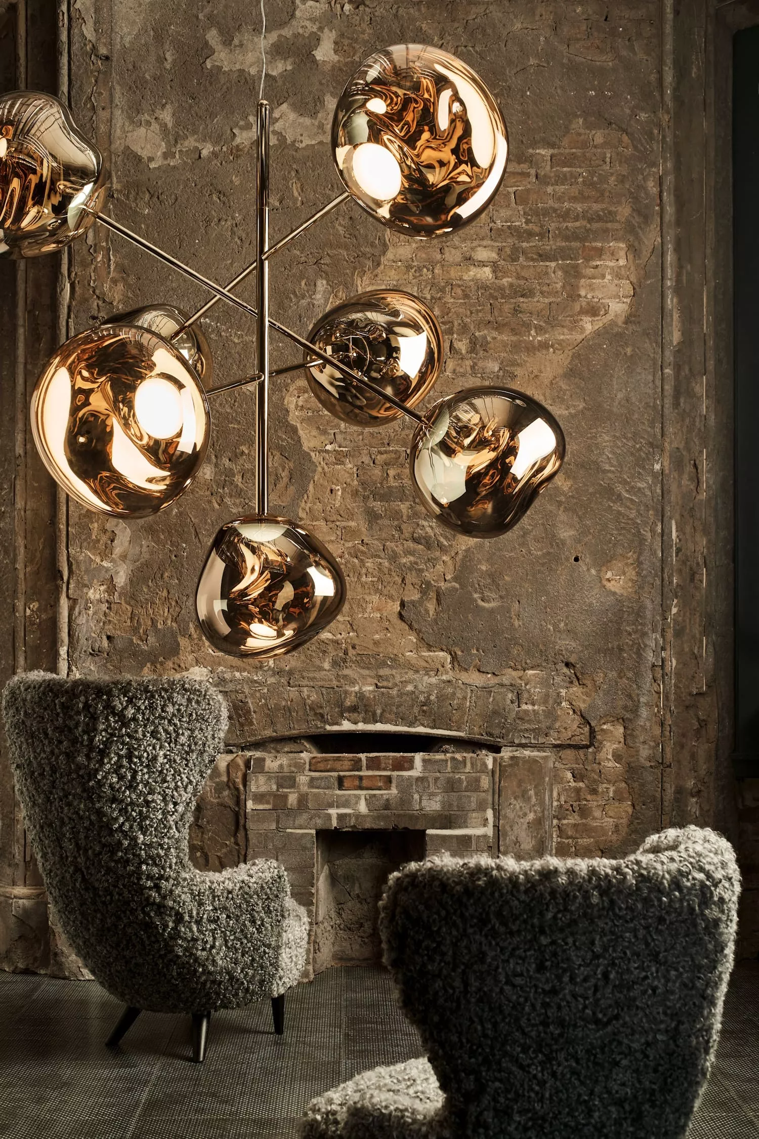Tom Dixon Kronleuchter GOLD MELT in einem rustikalen Raum mit Backsteinwand und gemütlichen Sesseln im Vordergrund.