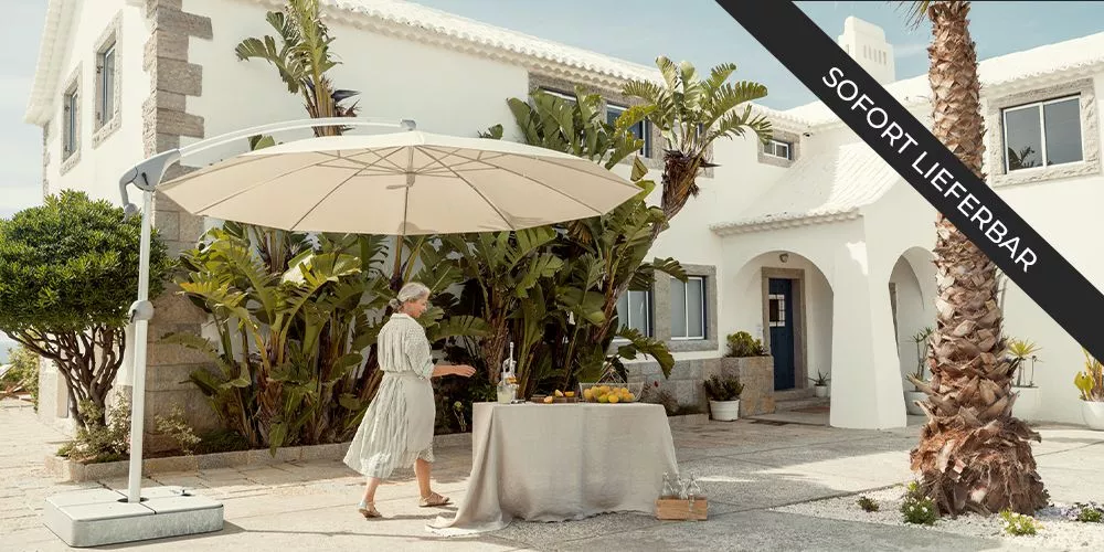 Glatz Sonnenschirm, cremefarben, auf einer Terrasse neben einem Tisch mit Obst und Getränken, vor einem weißen Haus mit Palmen.