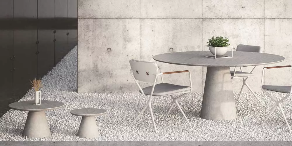 Royal Botania Conix Essgruppe in Grau auf Kiesboden vor Betonwand, bestehend aus rundem Tisch und Stühlen mit Armlehnen, stimmungsvolles Ambiente.