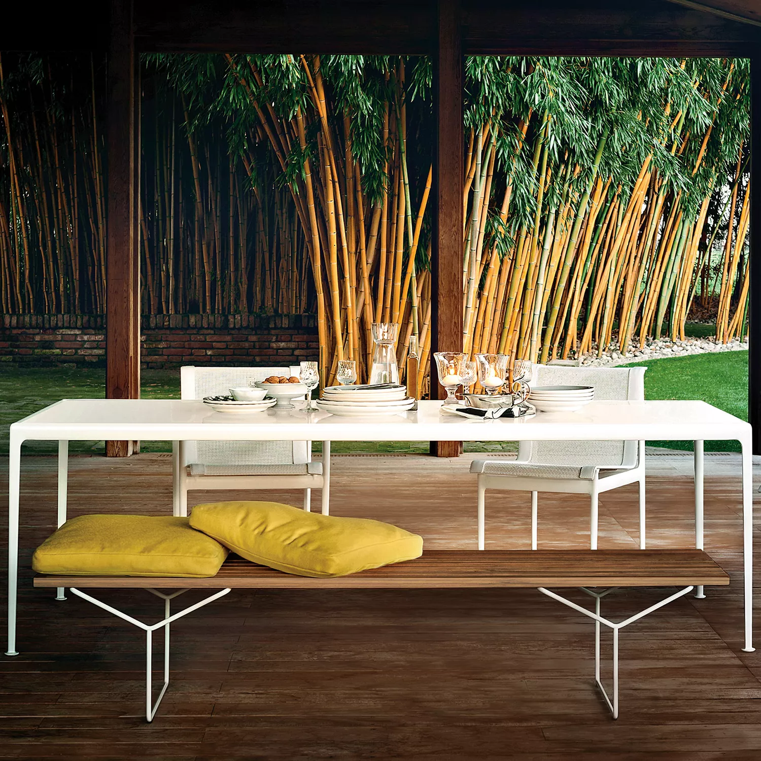 Knoll Harry Bertoia Lattenbank Teak steht vor einem gedeckten Esstisch im Freien mit Bambus im Hintergrund und gelben Kissen auf der Bank.