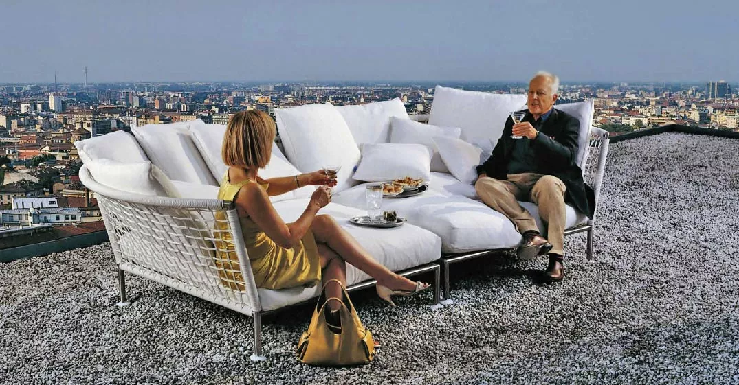 Eine Frau und ein Mann sitzen auf dem Coro Nest Sofa 120 cm abgerundet von Coro Italia auf einem Dach und genießen Getränke und Snacks.