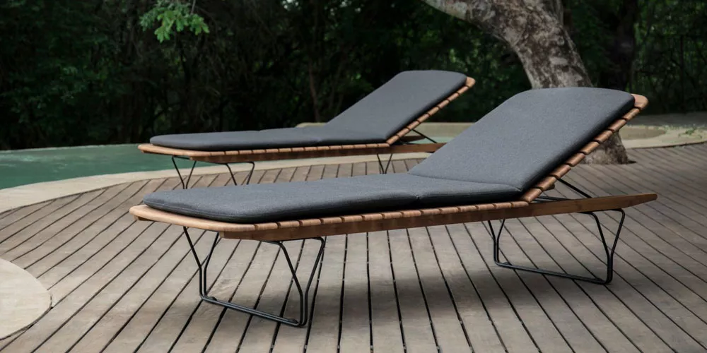 Zwei Houe Molo Sonnenliegen in Dunkelgrau auf einer Holzterrasse vor einem Pool, umgeben von grüner Natur.