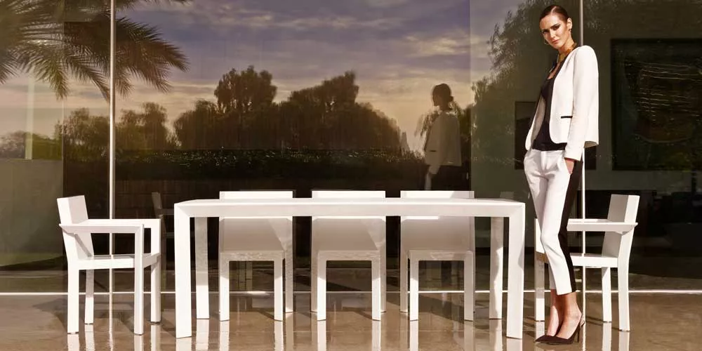 Frau in eleganter Kleidung steht neben der weißen Vondom Frame Essgruppe aus Kunststoff auf einer Terrasse bei Sonnenuntergang.