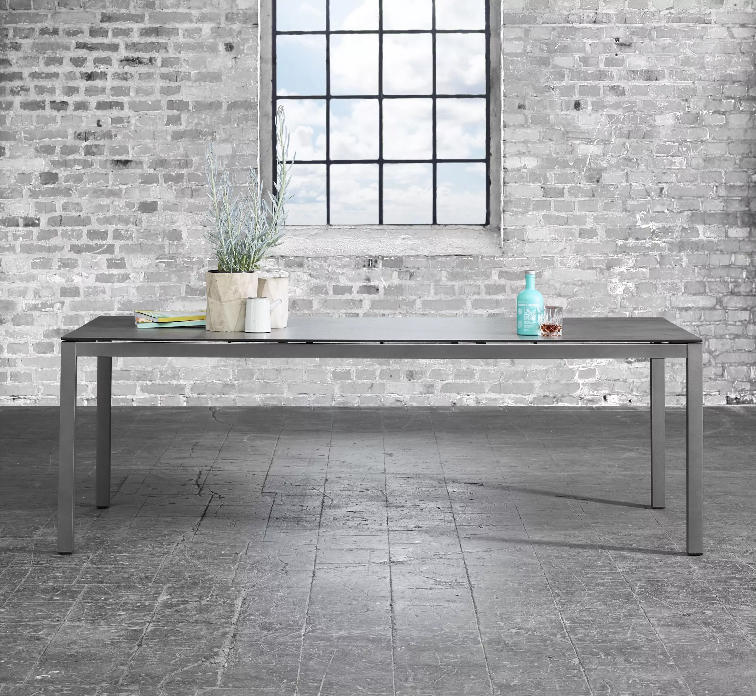 Solpuri Classic Alu Tisch in einem industriellen Ambiente mit einer Pflanze und einer Flasche auf der Tischplatte vor einer Backsteinwand und einem Fenster
