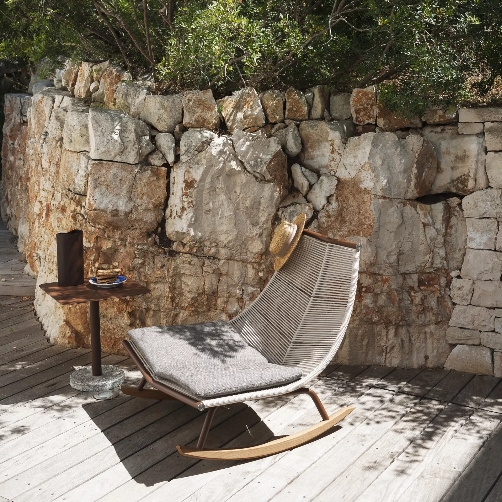 RODA Laze Kollektion: Beige Schaukelstuhl mit grauem Kissen auf Holzterrasse vor Steinmauer, sommerliche Gartenatmosphäre.