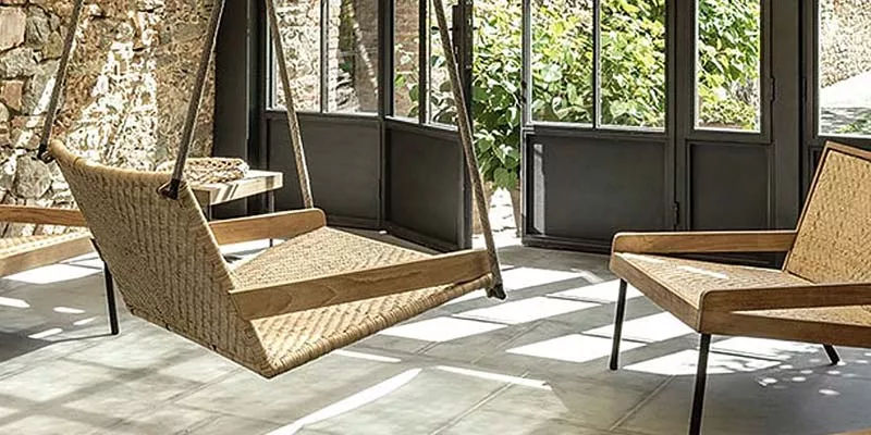 Hängesessel und Stuhl Ethimo Allaperto Verdana in Naturtönen auf einer sonnigen Terrasse mit Steinmauer und großen Fenstern.